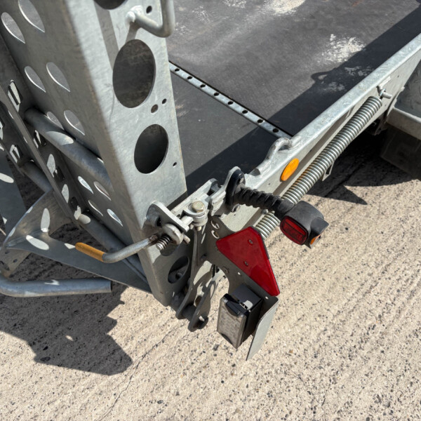 Close-up of a metal utility trailer rear corner showing perforated beams, a coiled suspension spring, and red taillight lenses on a gravel surface.