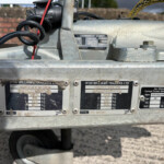 Close-up of a galvanized trailer hitch area with identification plates and a coiled black cable, on a gravel surface.