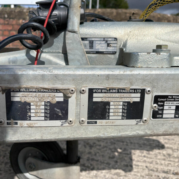 Close-up of a galvanized trailer hitch area with identification plates and a coiled black cable, on a gravel surface.