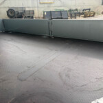 Empty flatbed trailer with tall metal sidewalls, textured floor, at a construction yard with equipment and gravel pile in the background.