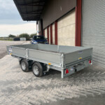 Silver utility trailer attached to a blue Isuzu pickup parked beside a corrugated metal warehouse.