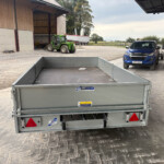 Metal flatbed trailer in the foreground at a farmyard, with a blue Isuzu pickup and a green tractor near a barn in the background