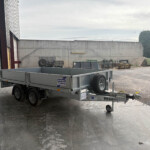 Silver flatbed trailer in a gravel yard beside a warehouse, with a concrete wall and scattered equipment behind it.