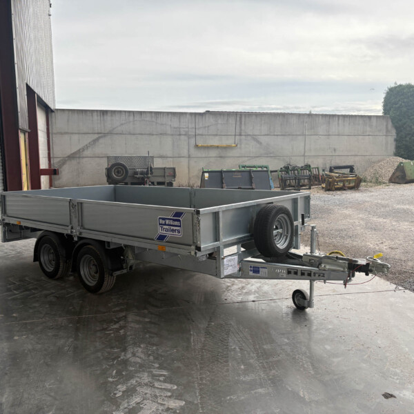 Silver flatbed trailer in a gravel yard beside a warehouse, with a concrete wall and scattered equipment behind it.