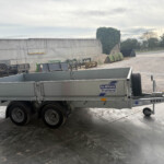 Silver Ifor Williams trailer in a gravel yard with farm equipment nearby and an overcast sky in the background.