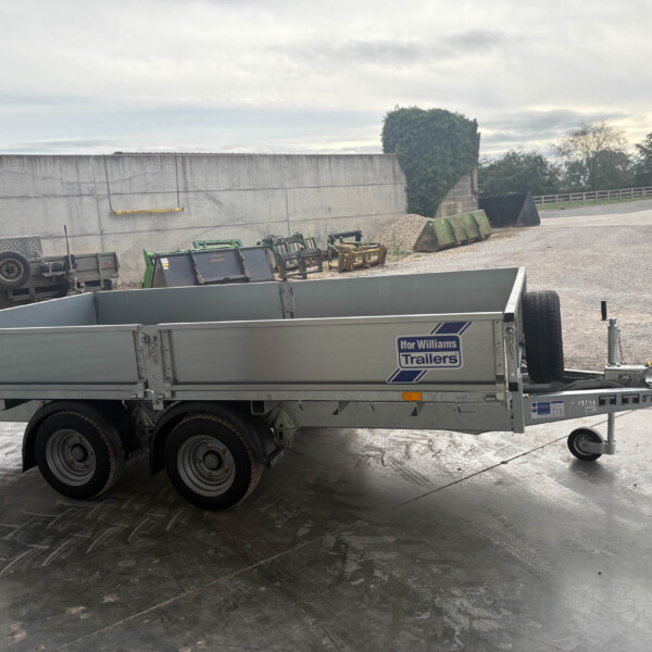 Silver Ifor Williams trailer in a gravel yard with farm equipment nearby and an overcast sky in the background.