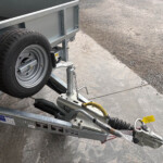 Close-up of a metal trailer frame with a mounted spare tire, hitch, and safety chains beside a gravel lot.