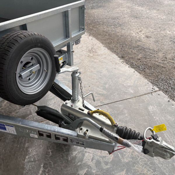 Close-up of a metal trailer frame with a mounted spare tire, hitch, and safety chains beside a gravel lot.