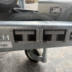 Two close-up data plates on a metal trailer frame reading 'IFOR WILLIAMS TRAILERS LTD' with a wheel visible below and a spare tire nearby.