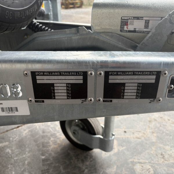 Two close-up data plates on a metal trailer frame reading 'IFOR WILLIAMS TRAILERS LTD' with a wheel visible below and a spare tire nearby.