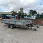 Wide view of a farm equipment lot with a large silver trailer in the foreground and a blue tractor beside a brick building with a Russell's sign in the background.
