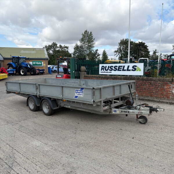 Wide view of a farm equipment lot with a large silver trailer in the foreground and a blue tractor beside a brick building with a Russell's sign in the background.