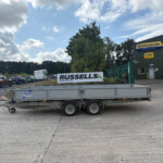 A flatbed trailer in the foreground with a parking lot of cars behind it and a New Holland Agriculture building to the right. A Russell's sign is visible in the midground under a cloudy sky.