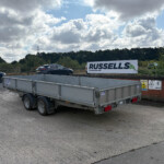 Grey flatbed trailer in a parking lot with a Russell's advertisement sign in the background; blue sky and clouds overhead.