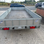 Rear view of a metal flatbed trailer in a parking area, with red taillights and branding stickers visible, cars and a brick wall in the background.