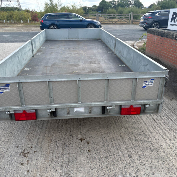 Rear view of a metal flatbed trailer in a parking area, with red taillights and branding stickers visible, cars and a brick wall in the background.