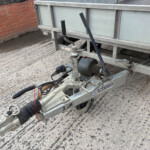 Close-up of a trailer hitch and coupling on a metal trailer frame beside a brick wall.