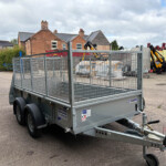Metal utility trailer with mesh sides in a paved yard, loaded with bags and equipment, brick houses in the background behind a fence.