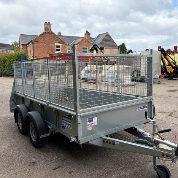 Metal utility trailer with mesh sides in a paved yard, loaded with bags and equipment, brick houses in the background behind a fence.