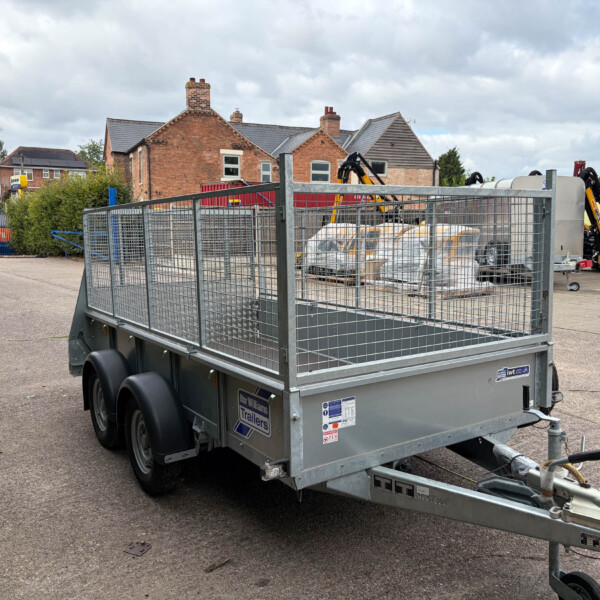 Metal flatbed trailer with mesh sides and double-axle wheels parked on a paved lot; construction equipment and brick houses in the background.