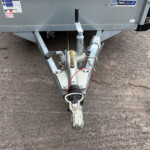 Close-up of a trailer hitch assembly on a gray trailer, showing the coupler, jack handle, safety chain, and wiring on a gravel surface.