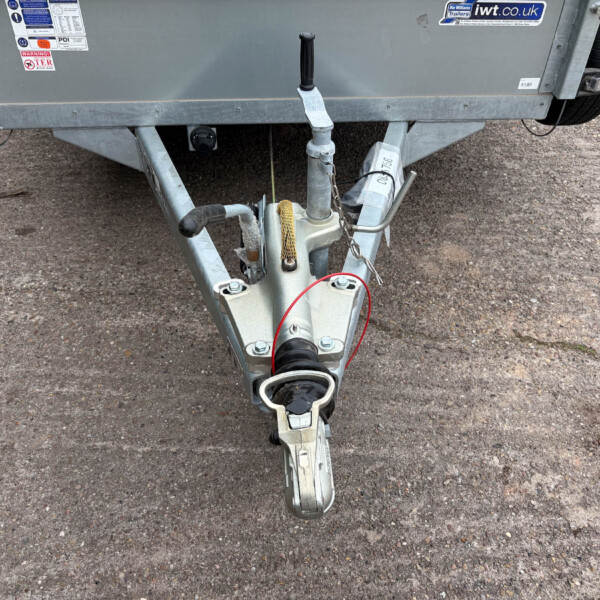 Close-up of a trailer hitch assembly on a gray trailer, showing the coupler, jack handle, safety chain, and wiring on a gravel surface.