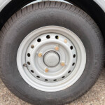 Close-up of a black tire mounted on a silver steel wheel with lug nuts on a rough pavement surface