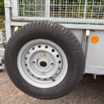 Close-up of a spare tire mounted on a trailer with a metal frame and grid sides, orange side marker visible on the trailer wall.