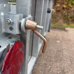 Close-up of a galvanized bolt latch on a gray metal frame, with a red reflective panel below and outdoor background.