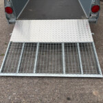 Open metal trailer ramp with a diamond-plate surface, ready for loading on a gravel driveway.
