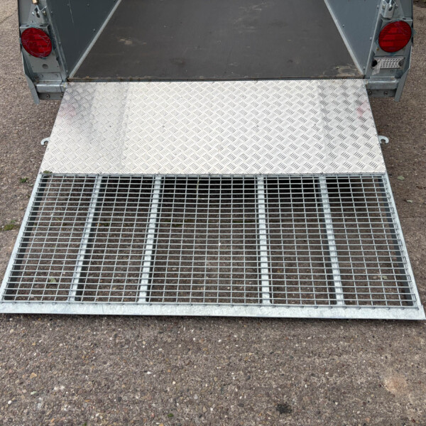 Open metal trailer ramp with a diamond-plate surface, ready for loading on a gravel driveway.