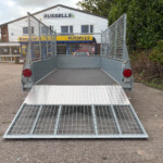 Metal utility trailer with a lowered diamond-plate ramp in a parking lot near a service shop with Russell's signage in the background.