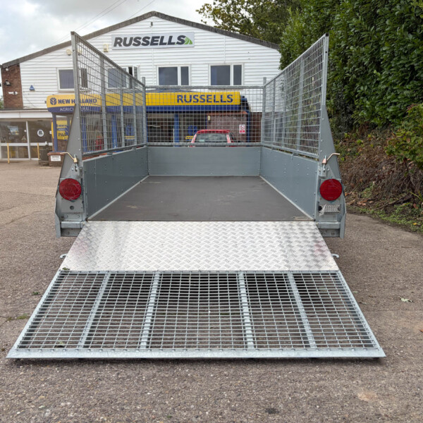 Metal utility trailer with a lowered diamond-plate ramp in a parking lot near a service shop with Russell's signage in the background.
