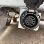 Close-up of a circular 7-pin trailer electrical connector with its cover open and mounting bracket visible under a vehicle.