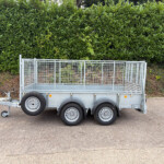 Metal utility trailer with tall wire mesh sides on a paved road, hedge behind it.