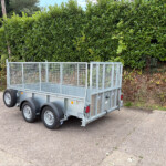 Metal utility trailer with tall mesh sides parked on a street in front of dense green hedges.