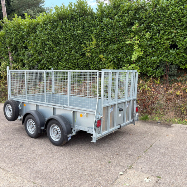 Metal utility trailer with tall mesh sides parked on a street in front of dense green hedges.