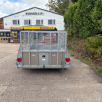 Rear view of a metal utility trailer in a parking lot, with a white building displaying Russell's and New Holland signs in the background.