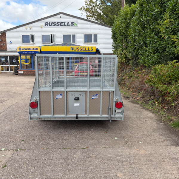 Rear view of a metal utility trailer in a parking lot, with a white building displaying Russell's and New Holland signs in the background.