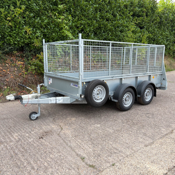Metal utility trailer with mesh sides parked on a paved area, with a tall green hedge in the background and a hitch at the front.