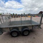 Metal-framed trailer in the foreground at an industrial yard, with yellow machinery and a service building in the background under a cloudy sky.
