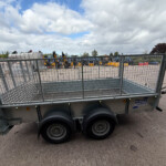 Metal utility trailer with mesh sides in a paved yard; construction machinery and a service building visible in the background under a cloudy sky.