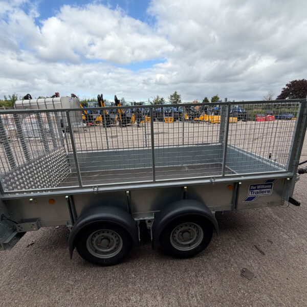Metal utility trailer with mesh sides in a paved yard; construction machinery and a service building visible in the background under a cloudy sky.