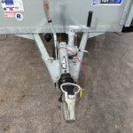 Rear view of a galvanized trailer hitch with a crank handle, safety chain, and spare tire against a concrete surface.