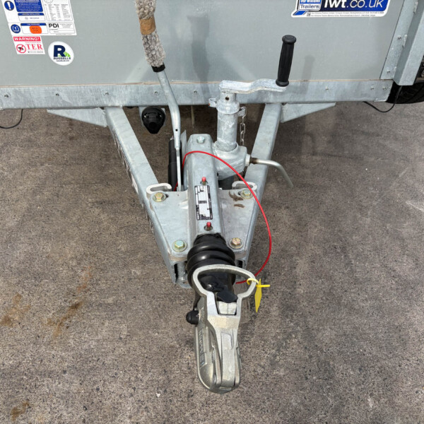 Rear view of a galvanized trailer hitch with a crank handle, safety chain, and spare tire against a concrete surface.