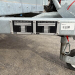 Close-up of a metal trailer frame with two weight plates labeled IFOR WILLIAMS TRAILERS LTD and capacity markings, plus part of a wheel and hitch visible in the foreground.