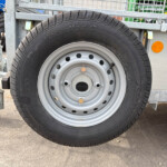 Close-up of a trailer wheel with black tire and silver rim, mounted on a metal trailer frame with a small step and orange side marker nearby.