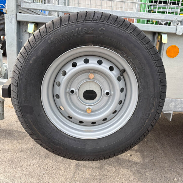Close-up of a trailer wheel with black tire and silver rim, mounted on a metal trailer frame with a small step and orange side marker nearby.