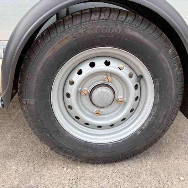 Close-up of a trailer tire mounted on a silver steel wheel with lug nuts and a gray fender above the wheel well.
