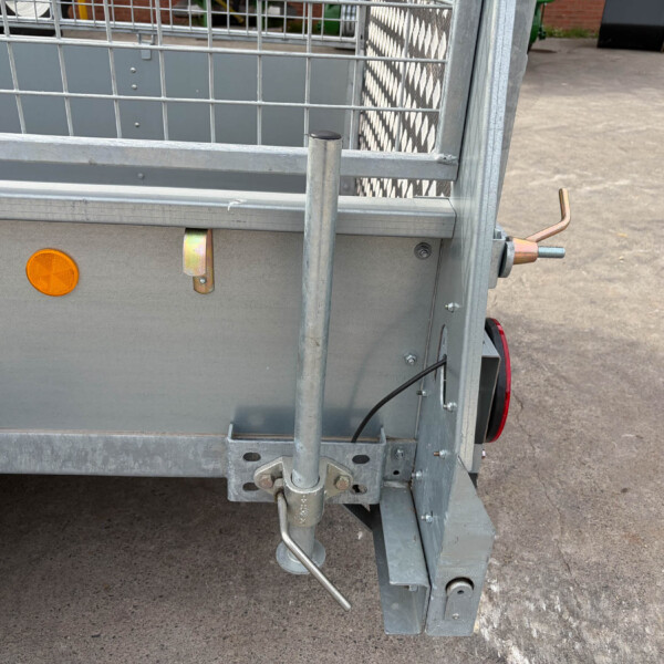 Rear corner of a metal utility trailer with a vertical support post, orange reflector, and hitch pin on a paved surface.
